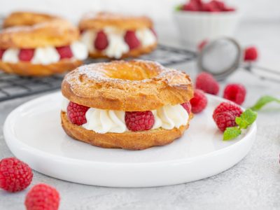 Choux pastries. Choux rings with cream of cream cheese or cottage cheese and fresh raspberries, dusted with powdered sugar on a white plate on a gray concrete background.